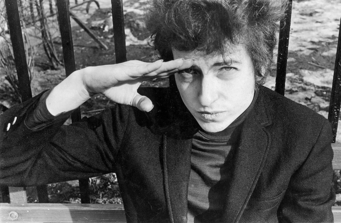 Bob Dylan, sitting on a bench in Christopher Park (across the street from the offices of the Village Voice since 1960), either salutes or shields his eyes from the sun, January 22, 1965<br/>